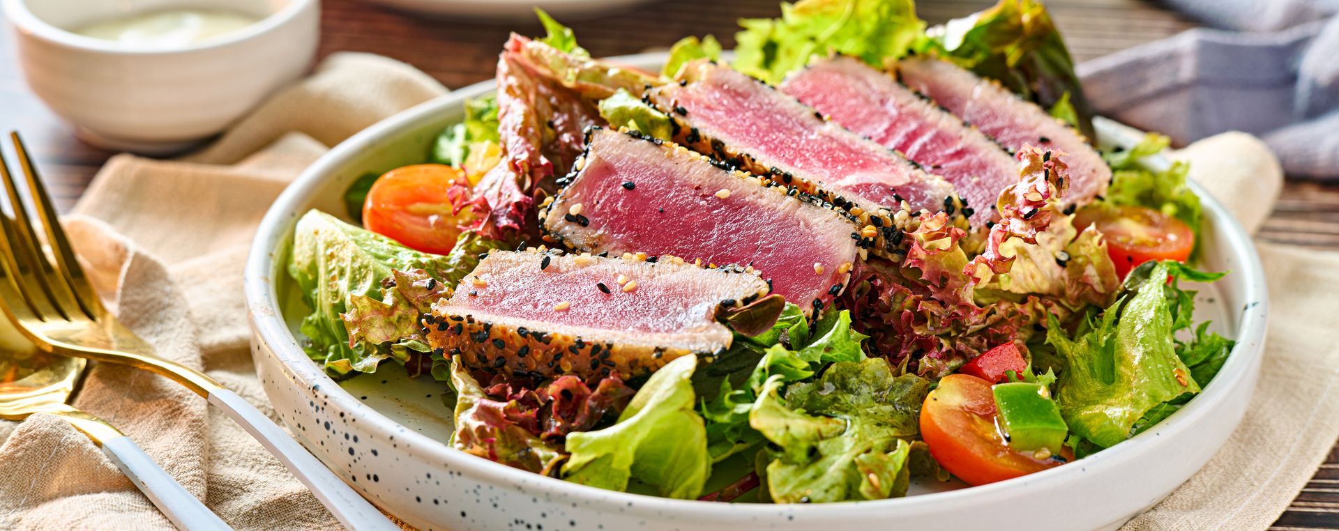 A seared ahi tuna salad with sliced fish, fresh greens, and cherry tomatoes in a shallow bowl with a side of dressing.