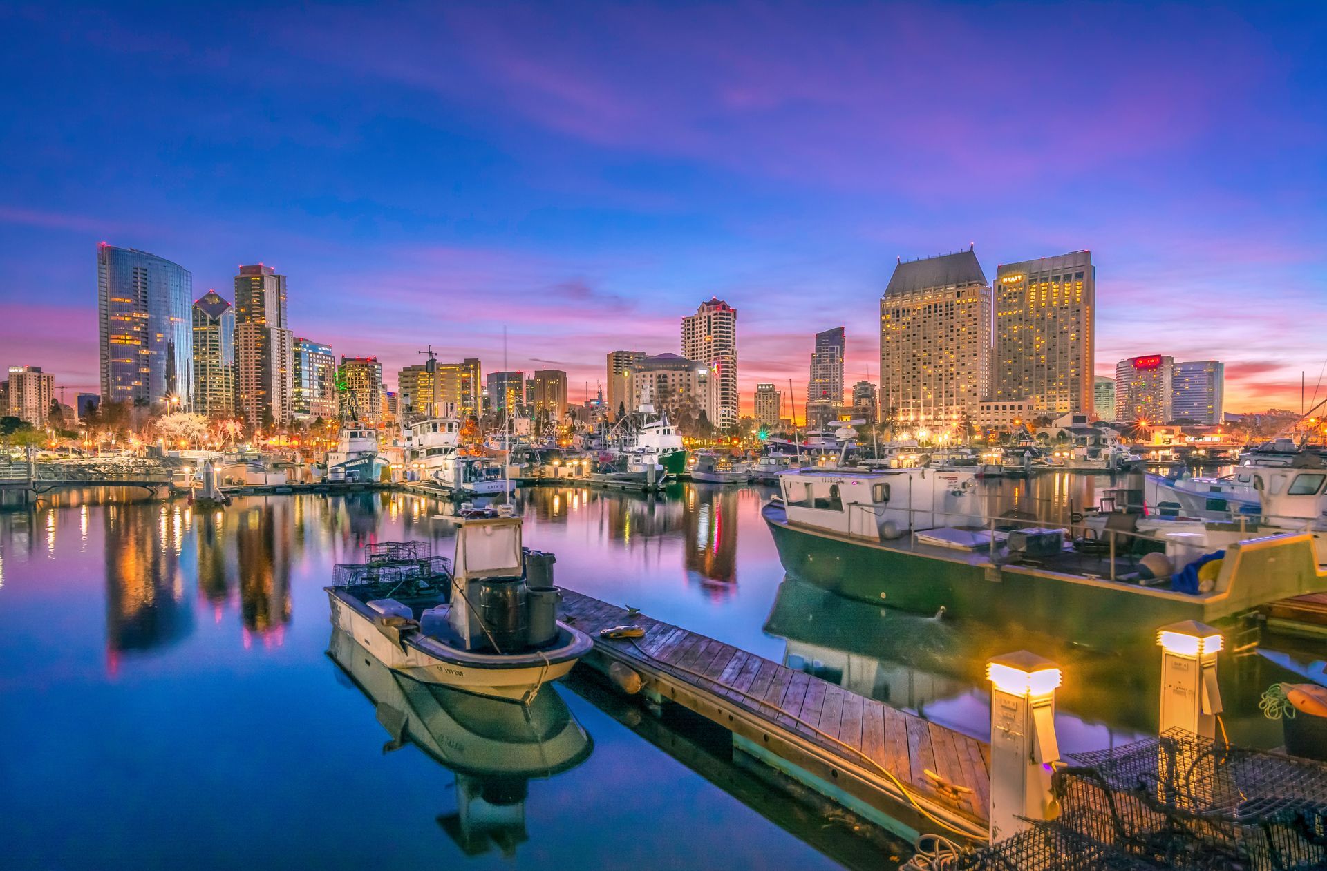 A vibrant sunset sky over the San Diego skyline, reflecting in the harbor waters beside docked boats and a wooden pier.