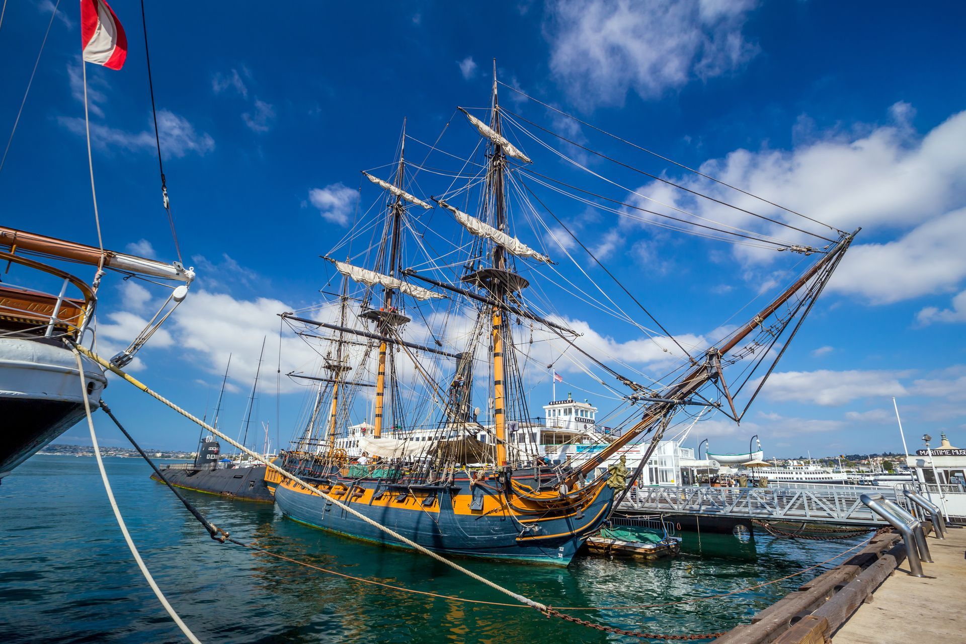 A tall, three-masted wooden sailing ship docked at a pier under a bright blue sky with scattered clouds.