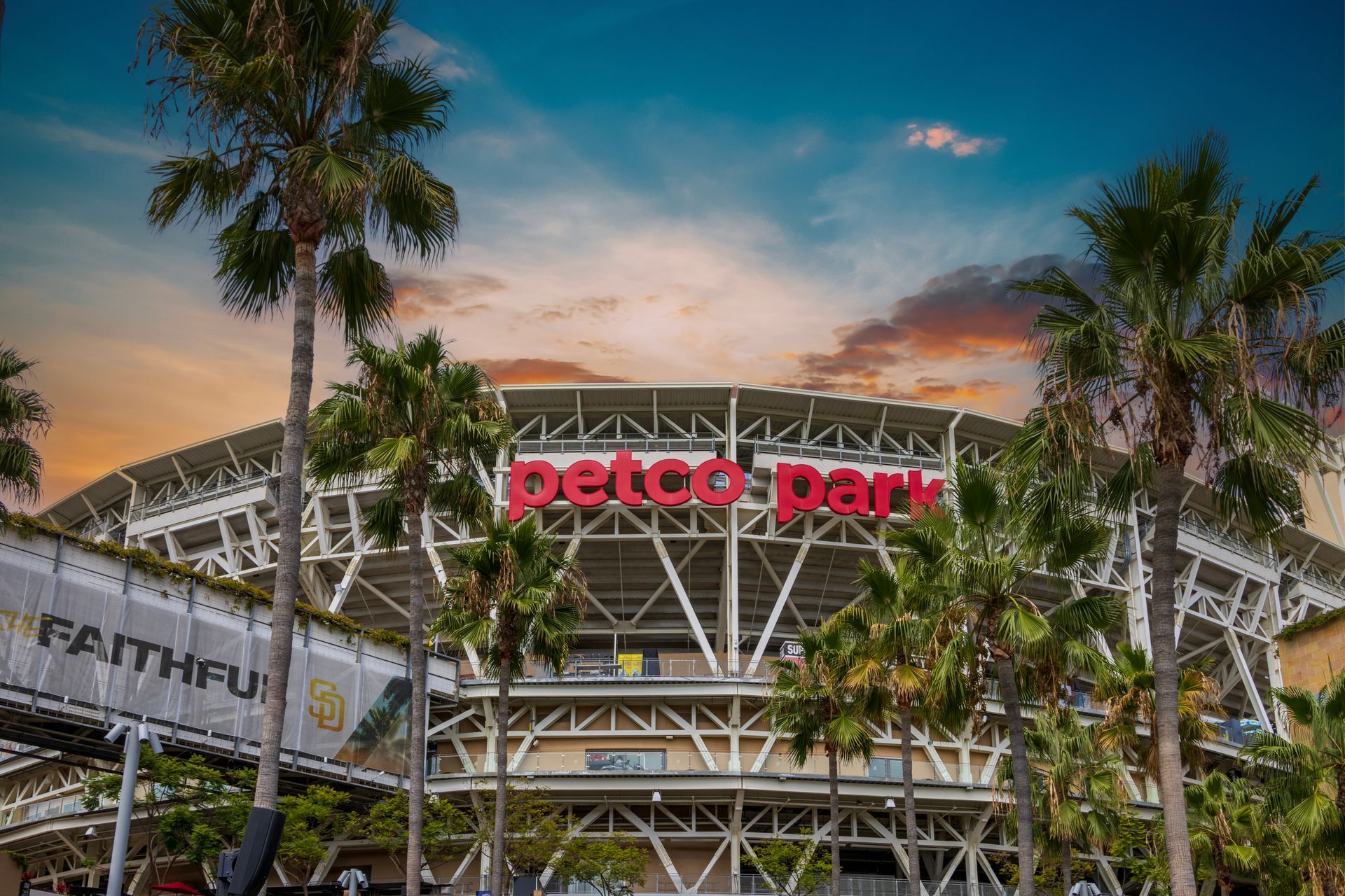 The exterior of Petco Park stadium in San Diego, featuring the red logo against a structural framework under a sunset sky.