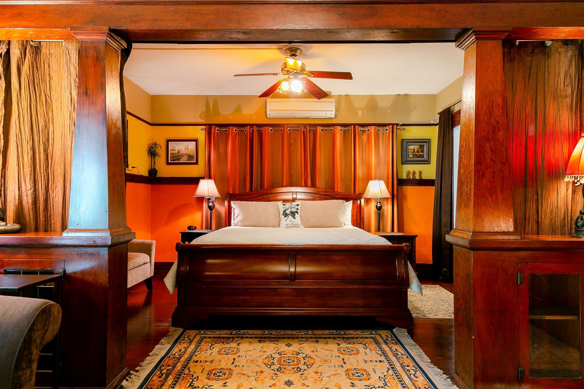 A bedroom with a dark wood sleigh bed, orange walls, floor-to-ceiling curtains, and an ornate patterned rug.