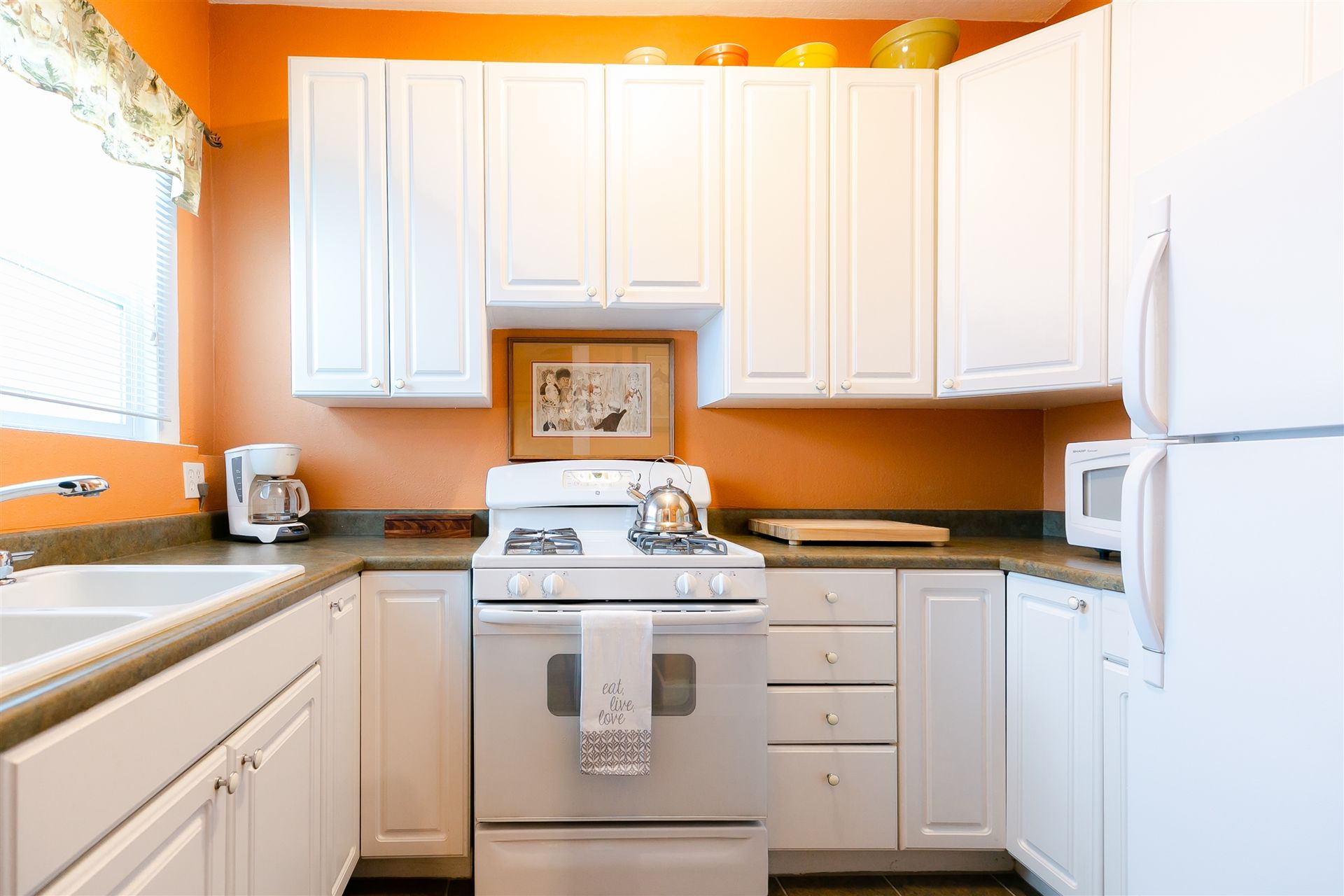 A kitchen with orange walls, white cabinets, white appliances, and a window with a patterned valance.