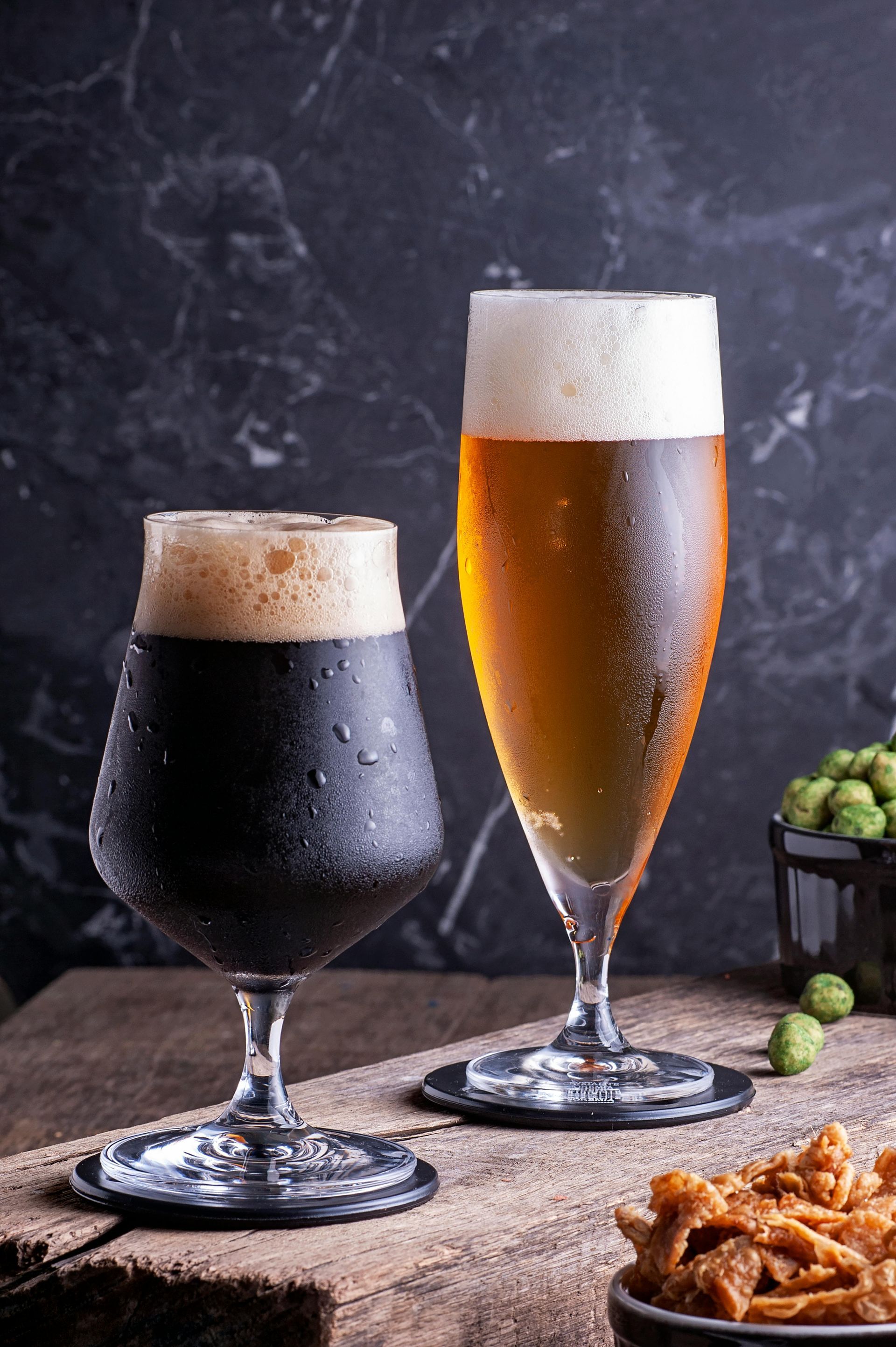 A dark stout and a light golden beer in stemmed glasses on a wooden surface, with a bowl of snacks nearby.