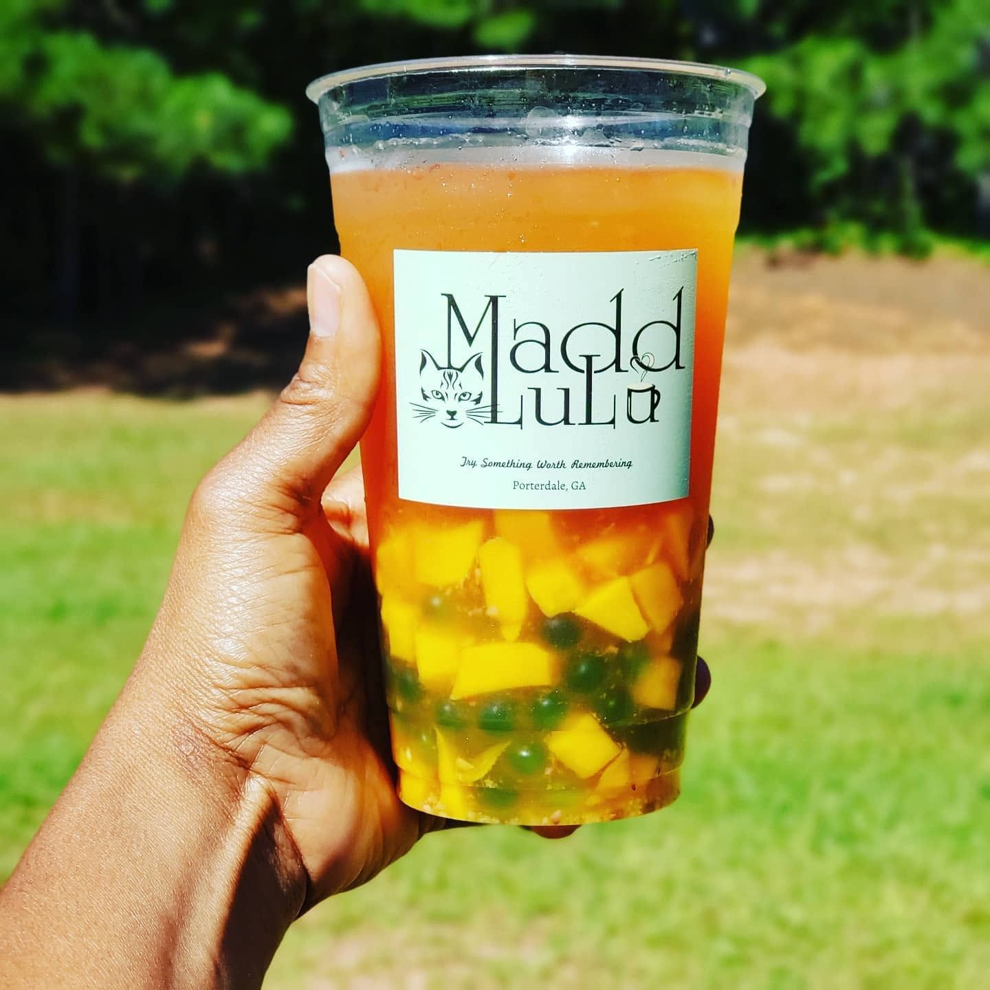 Hand holding a clear cup of orange drink with fruit pieces and a Madd LuLu label outdoors.