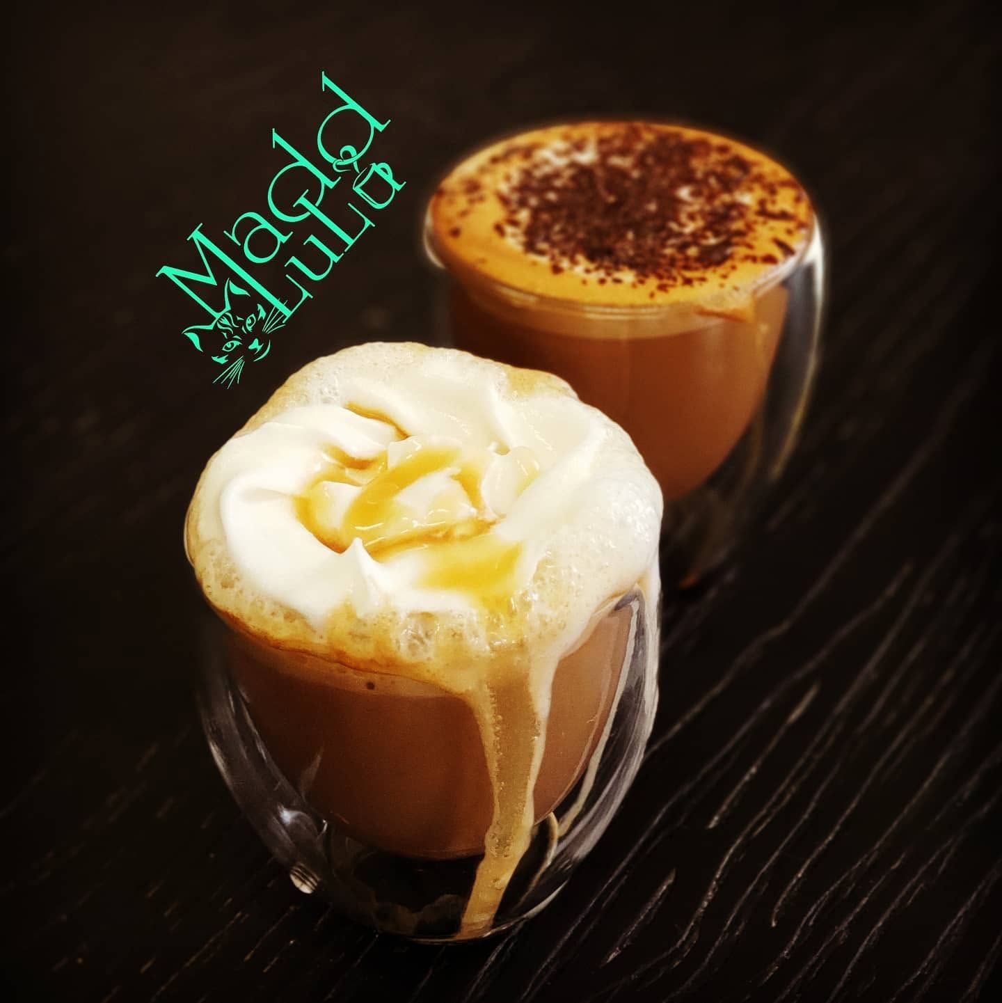 Two frothy caramel coffee drinks on a dark table, one topped with whipped cream and honey drizzle