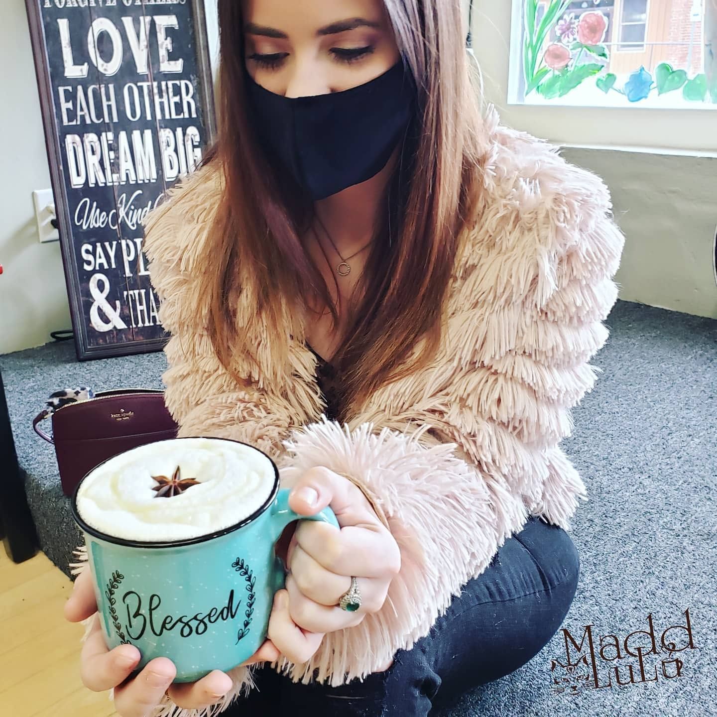 Woman in a black mask holding a teal “Blessed” mug of coffee indoors.