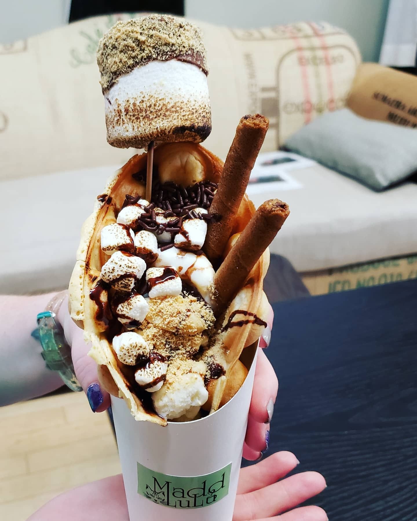 Chocolate dessert cone topped with marshmallows and wafer sticks, held indoors against a sofa background