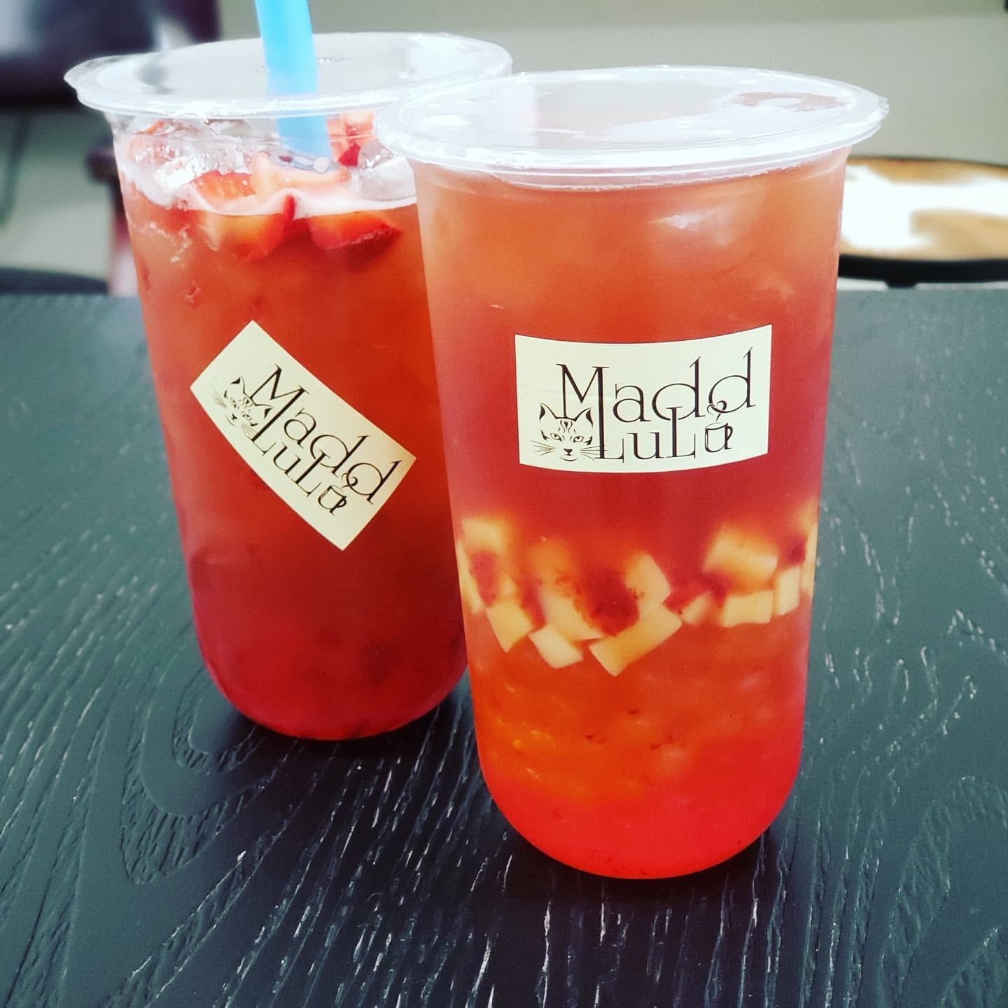 Two red fruit drinks in clear plastic cups with white lids, one with a blue straw and fruit pieces inside.
