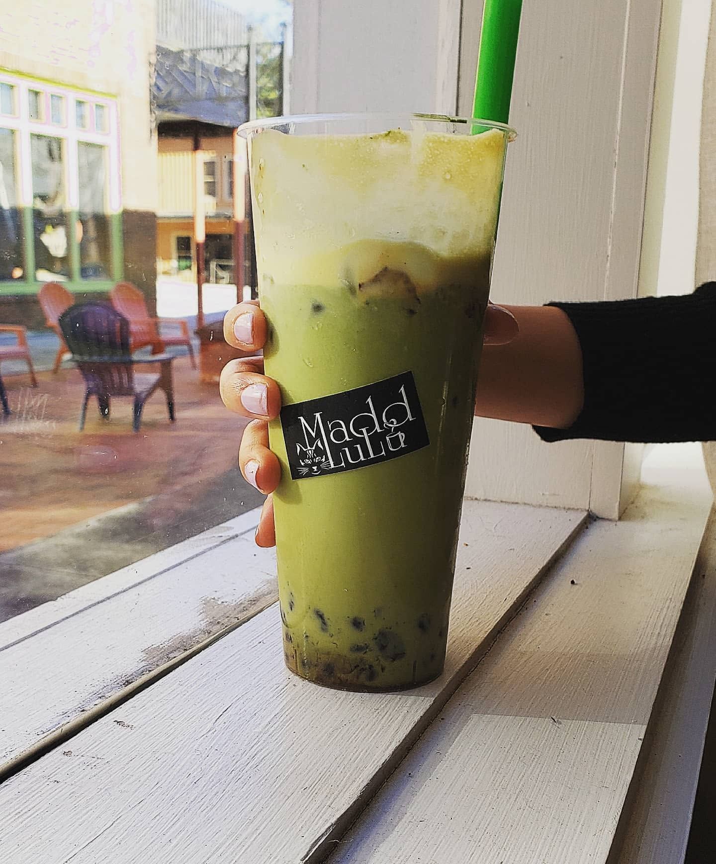 Tall green iced drink in a clear cup, held by a hand on a white windowsill.