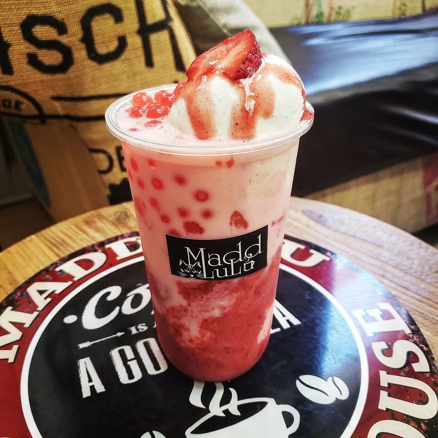 Strawberry whipped-cream drink in a clear cup on a tabletop, with red drizzle and a café logo.