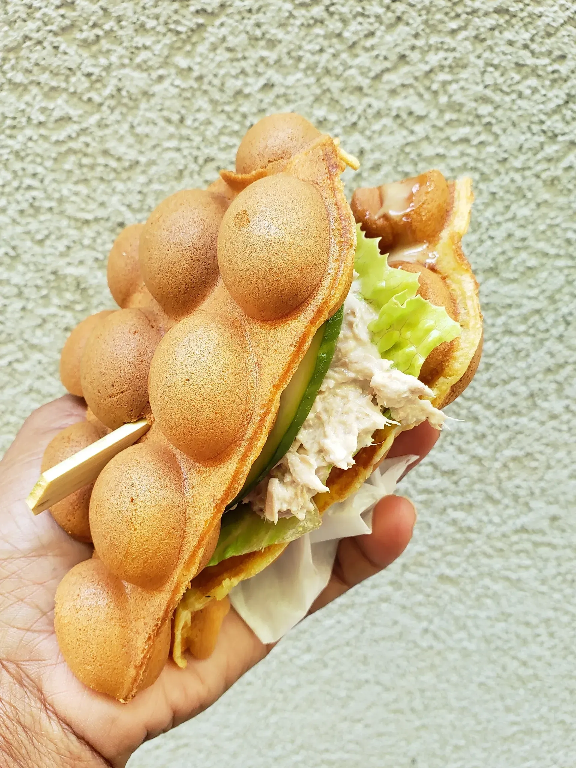 Hand holding a fish-shaped waffle sandwich with lettuce and filling against a textured wall