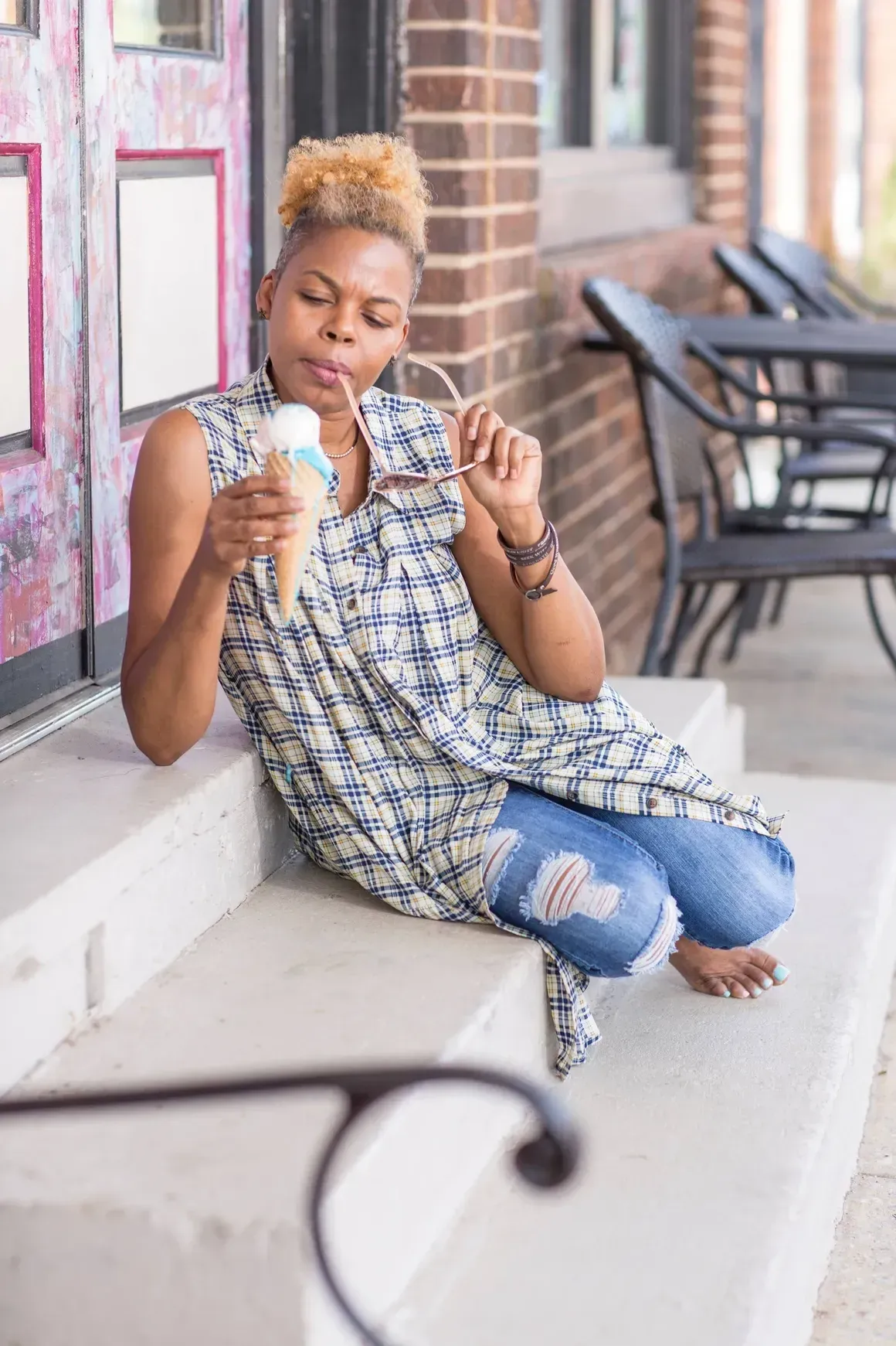 Person sitting on a porch bench eating ice cream cone, with empty café chairs in the background.