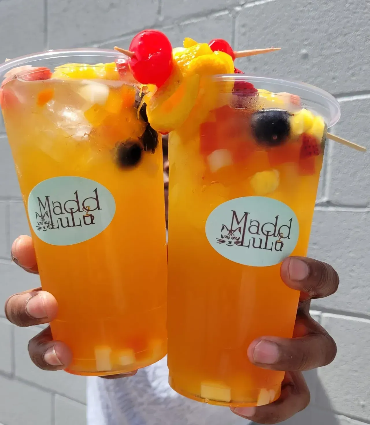 Two orange fruit drinks with ice, fruit garnish, and cherries held in hands
