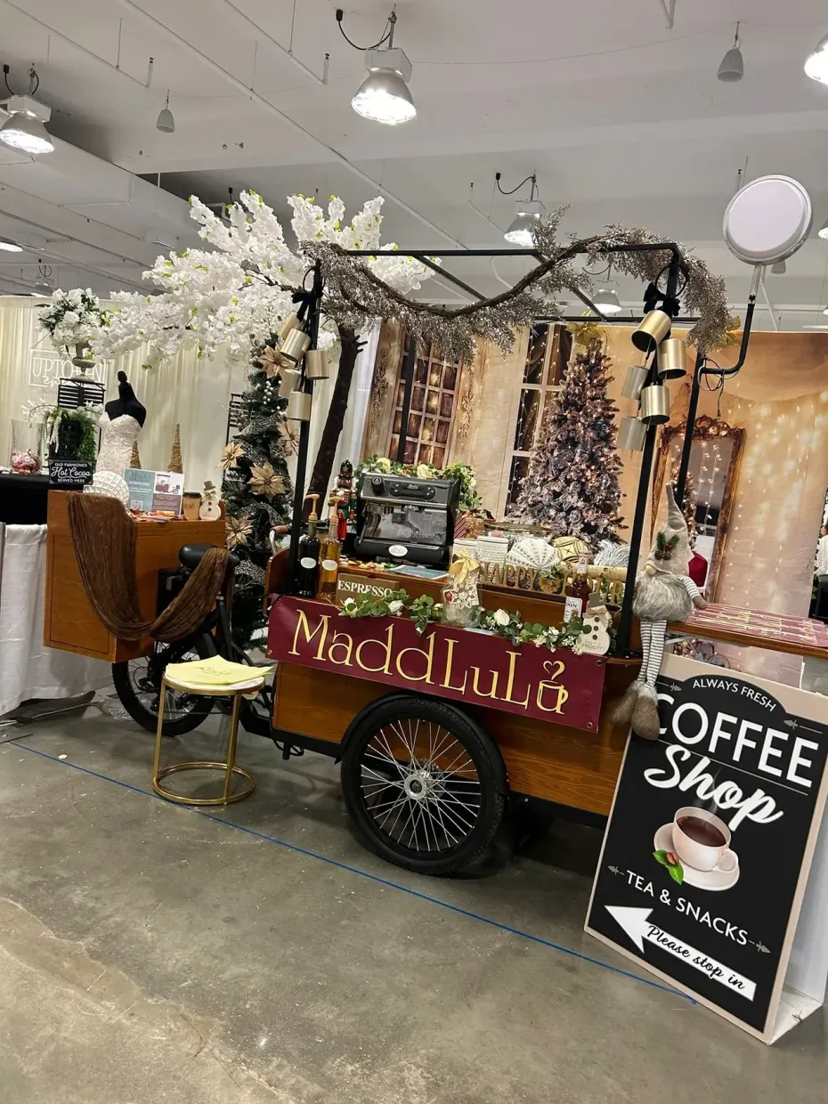 Rustic coffee cart booth with red sign, black wagon wheel, and white floral decor at an indoor market