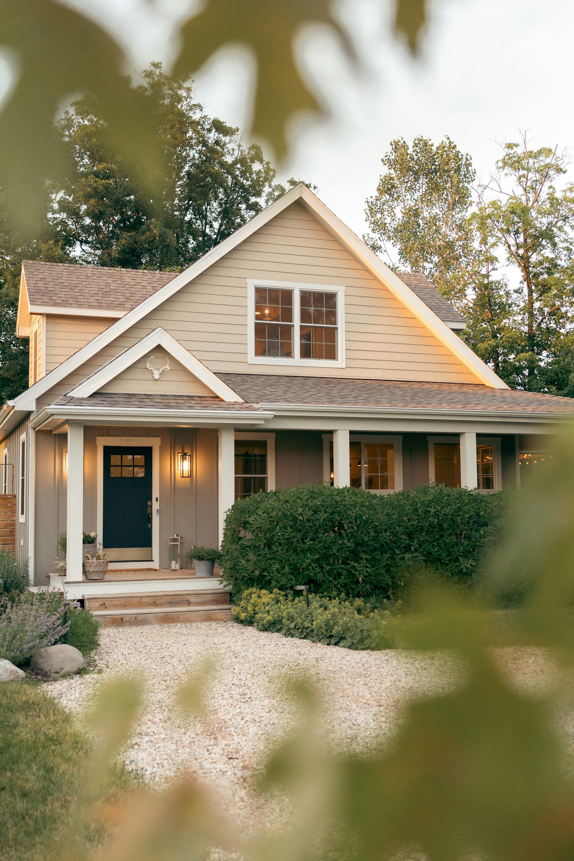 Cottage-style house with beige siding, dark blue door, and porch. Bushes and trees frame the front.