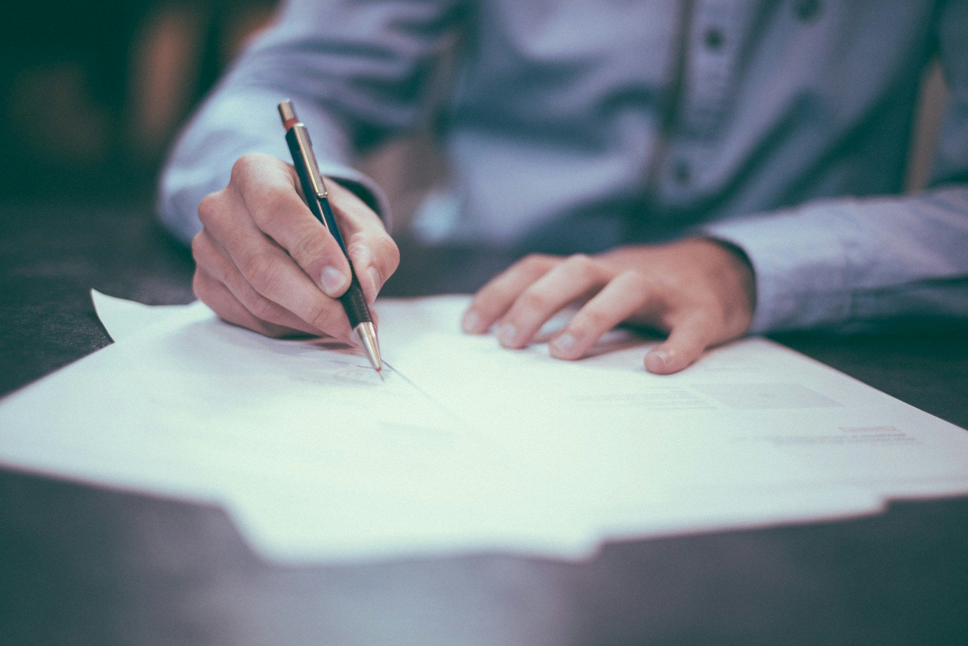 Person's hands writing on white paper with a pen, wearing a blue shirt.