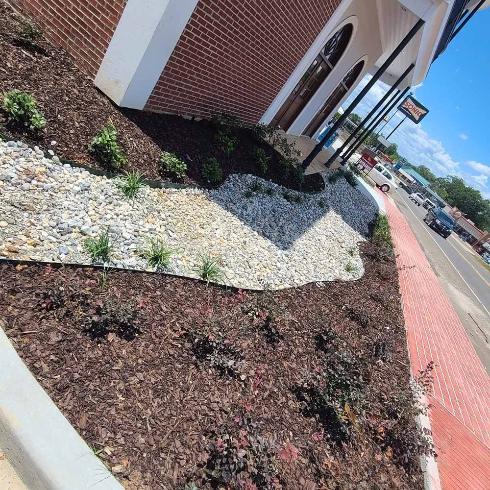 Landscaped area with brown mulch, gray rocks, green plants, and a brick building along a street.