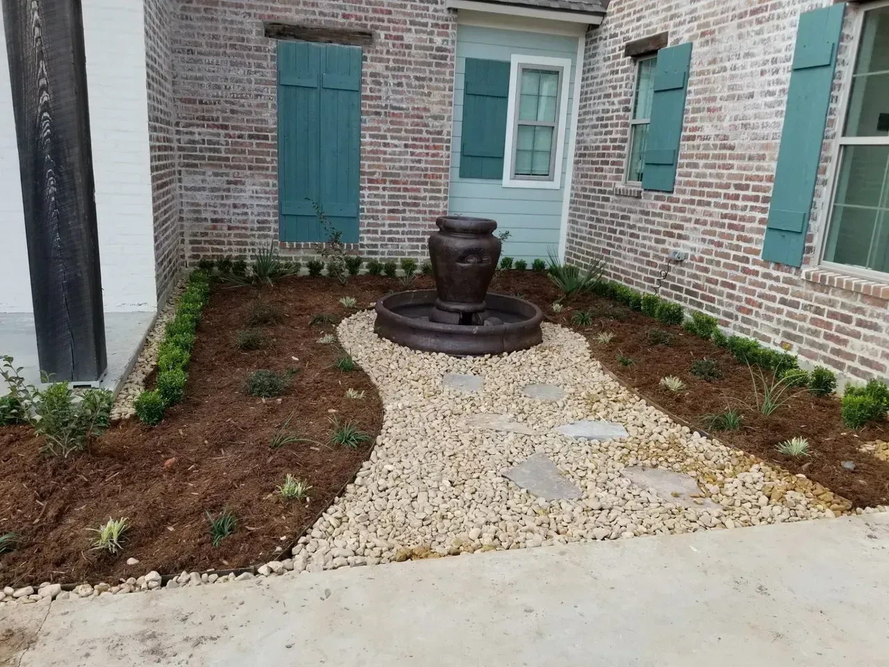 Brick building exterior with a fountain in a garden bed with pebbles and mulch.