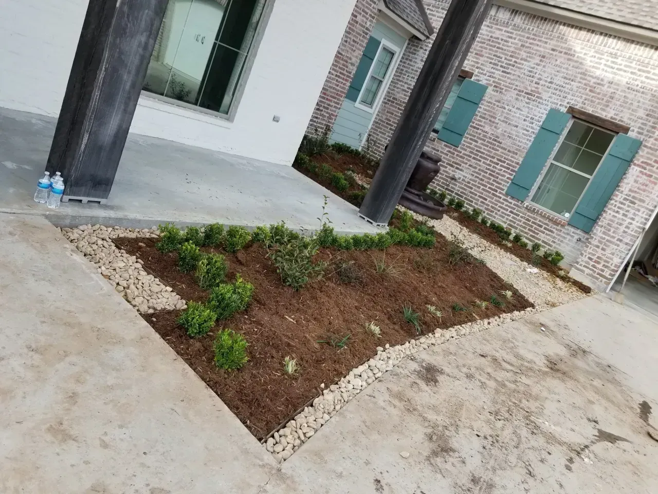 Small garden bed with mulch and shrubs, bordered by gravel. Located near a brick building with teal shutters.