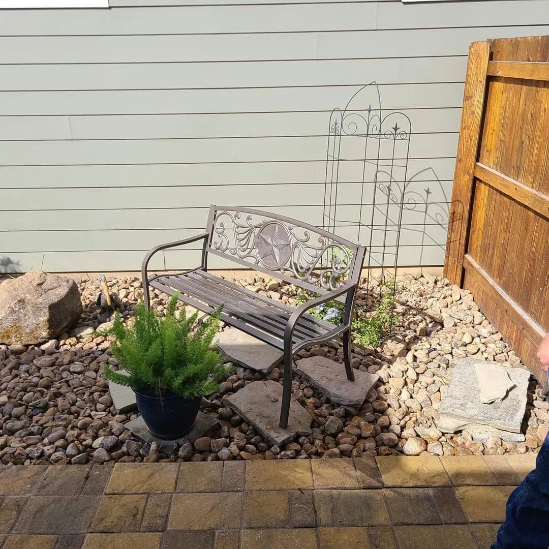 Metal bench in a backyard surrounded by rocks and a potted fern.  A wooden fence and white siding are in the background.