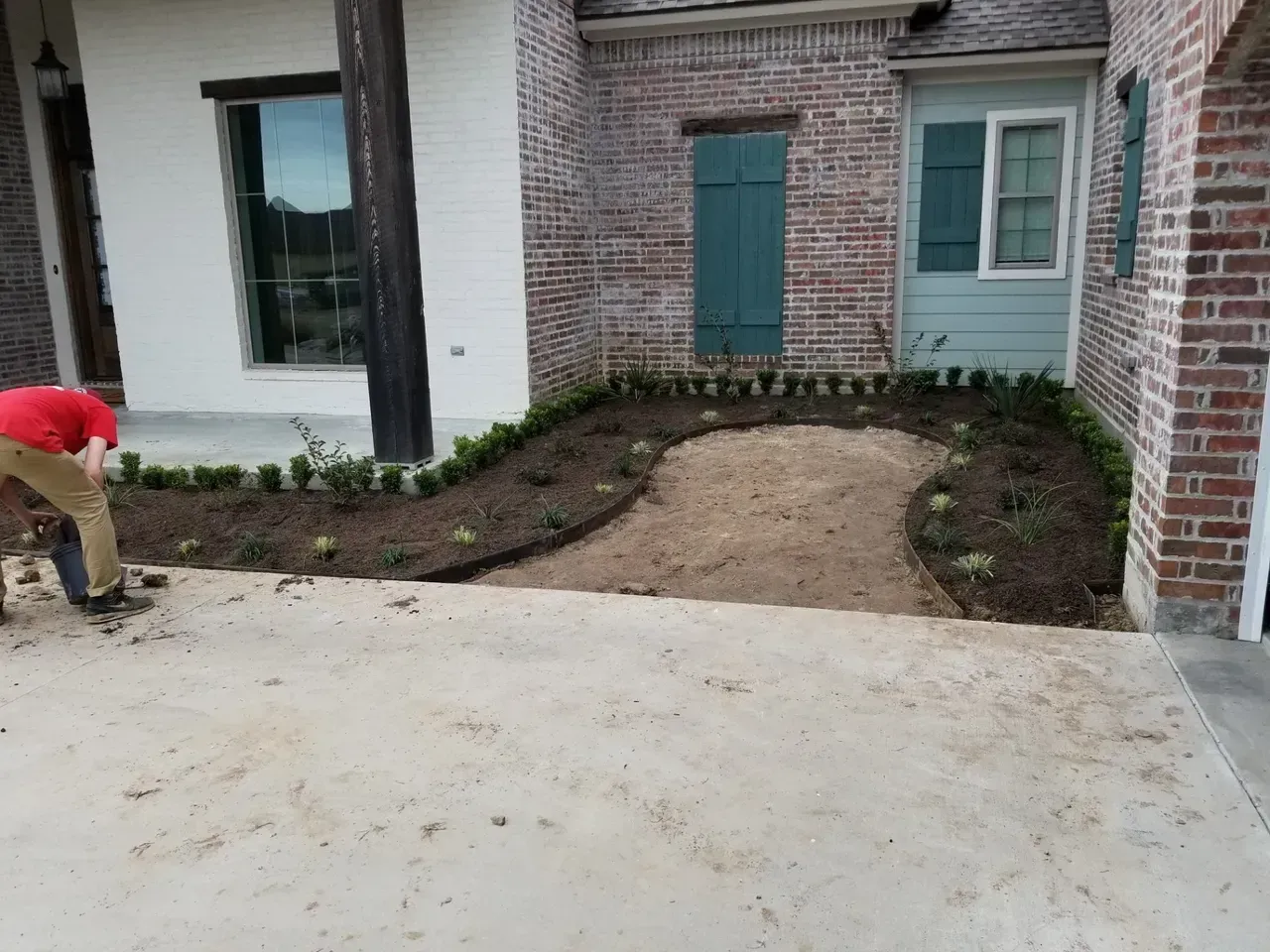 Man working on landscaping bed in front of brick building. Dirt pathway, newly planted greenery.