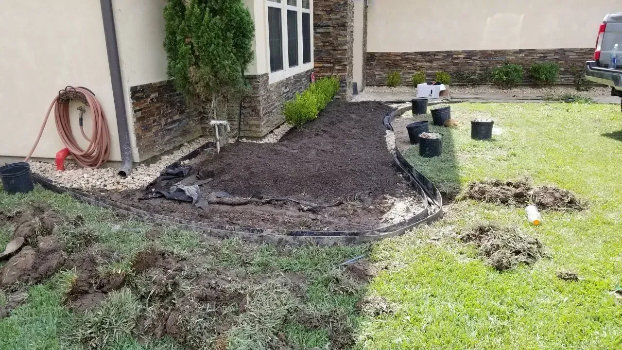 Newly planted garden bed with dark soil, surrounded by grass and a house. Black pots are nearby.