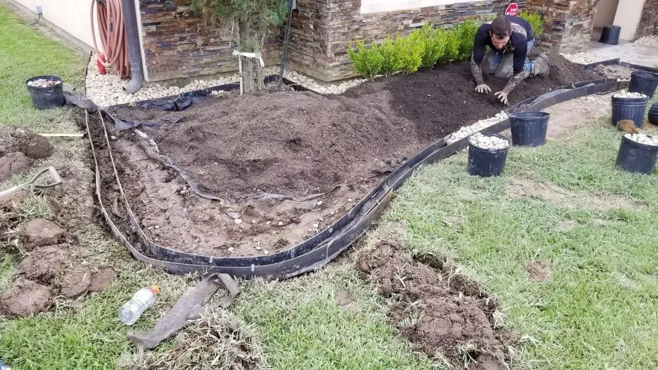 Man planting in a garden bed with black edging, surrounded by dirt, grass, and potted plants.