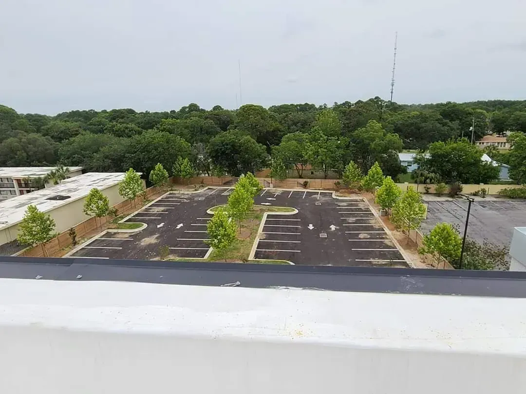 Empty parking lot surrounded by small trees, seen from above, with a treeline and cloudy sky in the background.