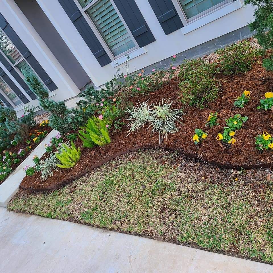 Flower bed with mulch and various plants, bordered by a green lawn, in front of a house.
