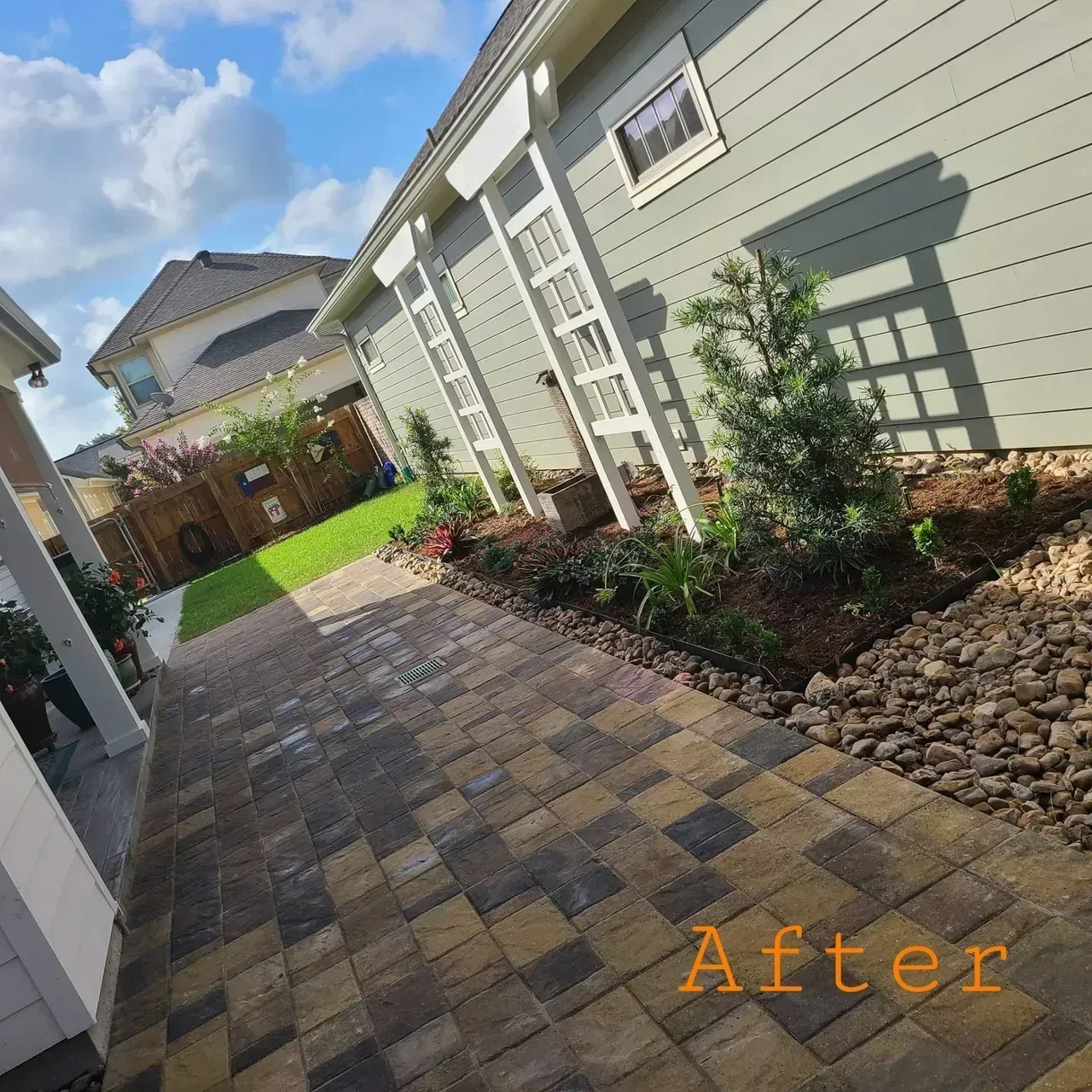 A paved walkway leads past a house with a landscaped garden. Bright blue sky overhead.