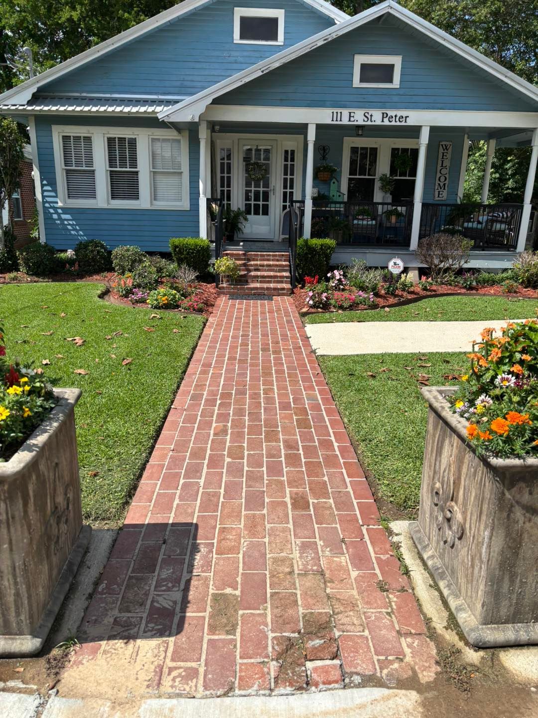 Brick pathway leading to a blue house with porch; flowerbeds and potted plants line the walkway.