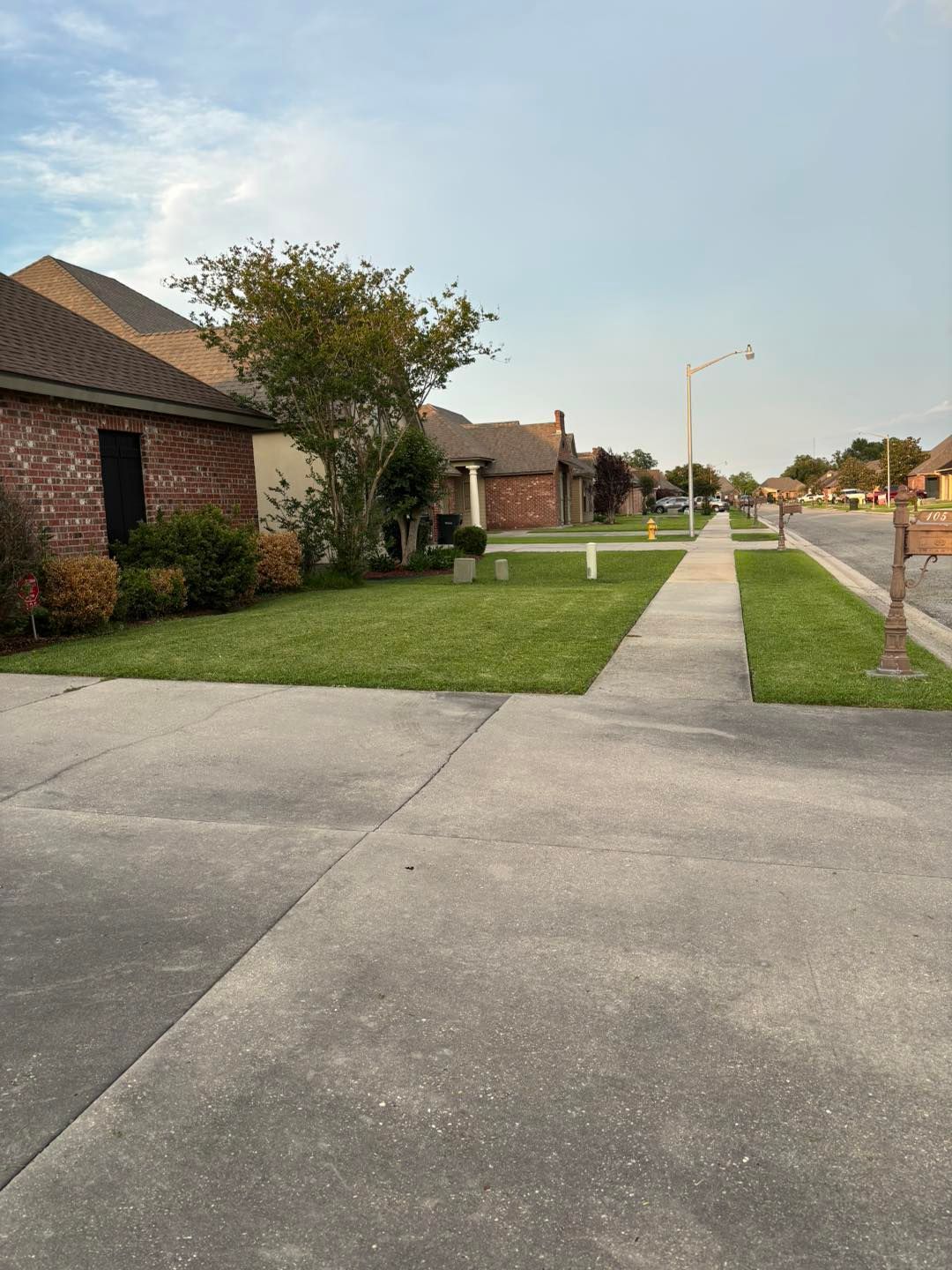 Suburban street scene with houses, sidewalk, and green lawns under a cloudy sky.