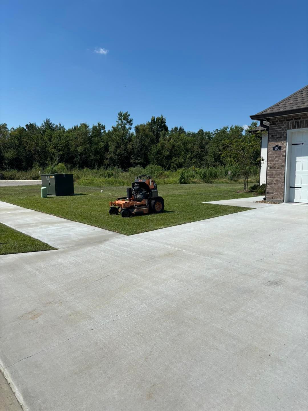 Lawnmower on a green lawn, near a house and a concrete driveway. Sunny day with blue sky and trees.