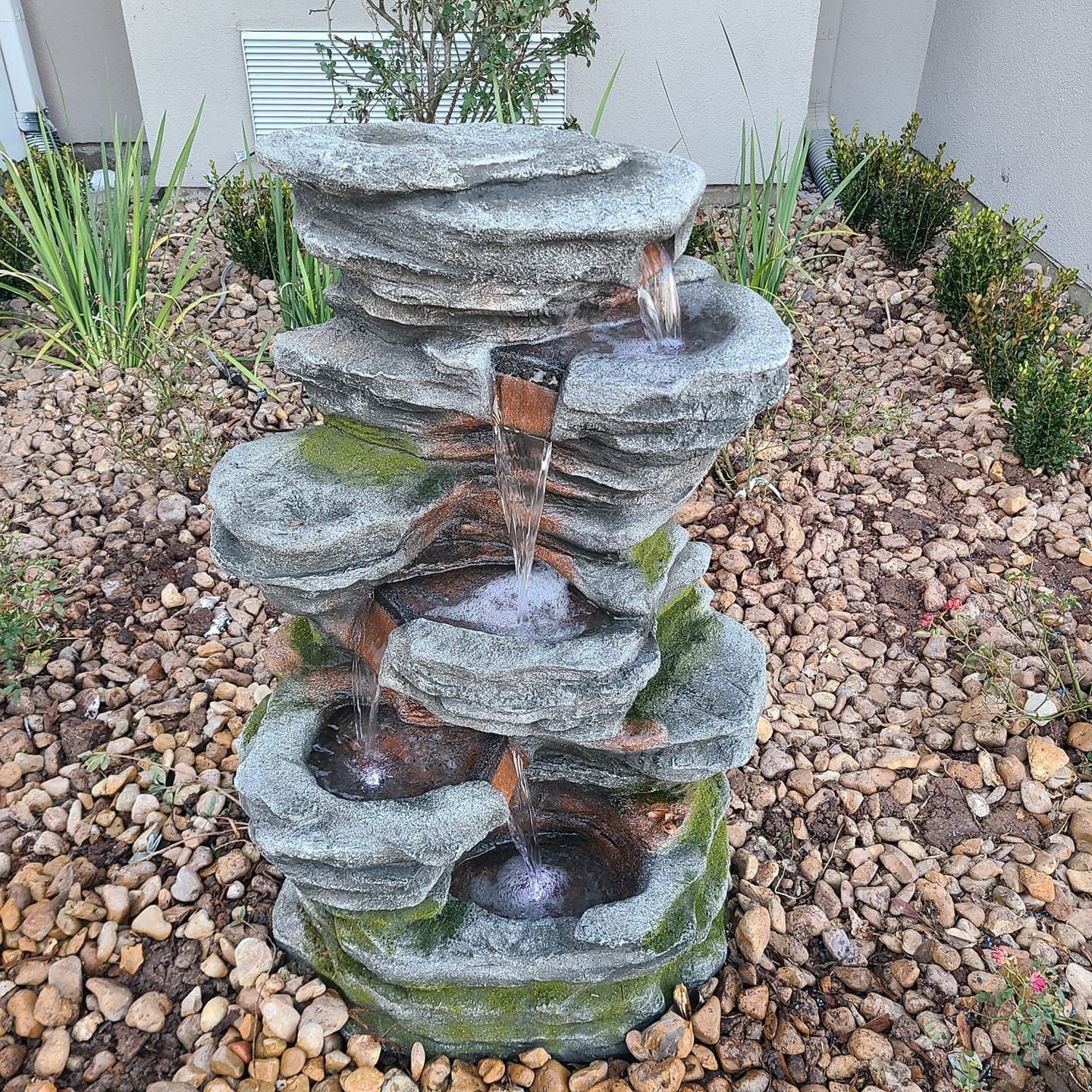 Rock fountain with water cascading over tiered gray stones, surrounded by small brown rocks and greenery.