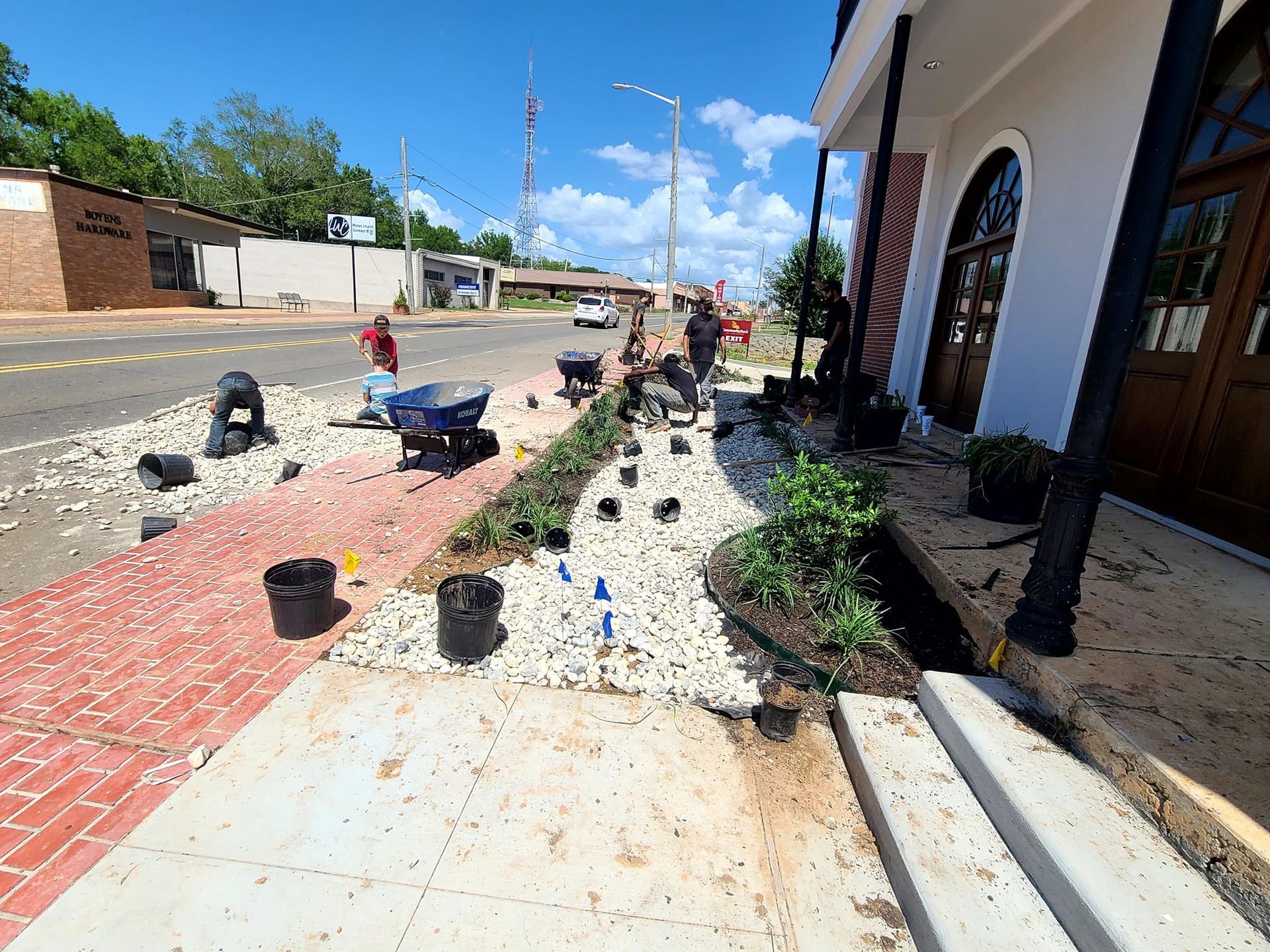 Workers planting greenery along a sidewalk. Gray stone, brick, and red paving stones are visible. Blue sky.