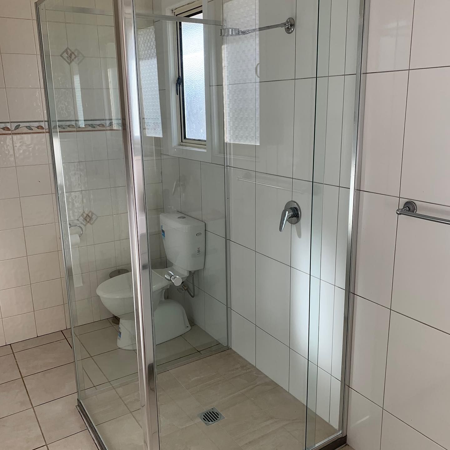 Shower stall with glass doors, toilet, and window in a small bathroom. Light-colored tiles on walls and floor. — Moore Park Beach Glass & Security in Elliott Heads, QLD