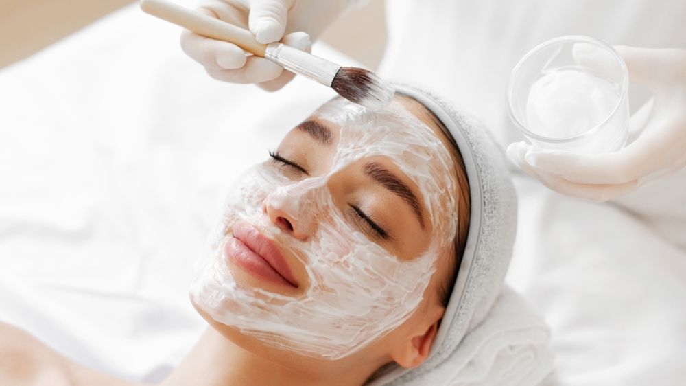 Facial spa treatment with cream applied to a woman's face by a brush, hands wearing gloves nearby.