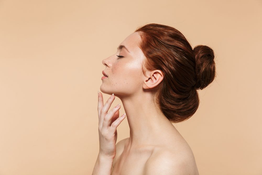 Profile of a person with hair in a bun, gently touching their chin against a beige background