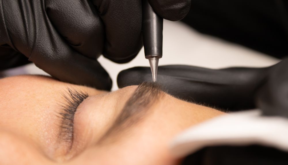 Gloved artist applying a cosmetic tattoo near a closed eye with a handheld tool