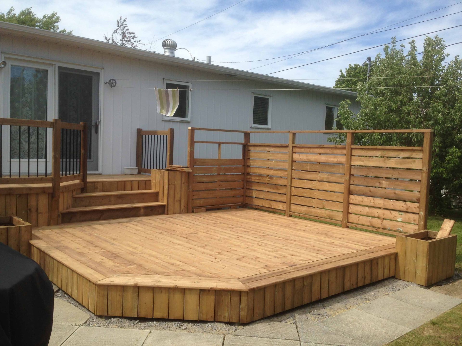 Terrasse en bois dans le jardin, avec marches, rambarde et brise-vue, à côté d'une maison blanche.