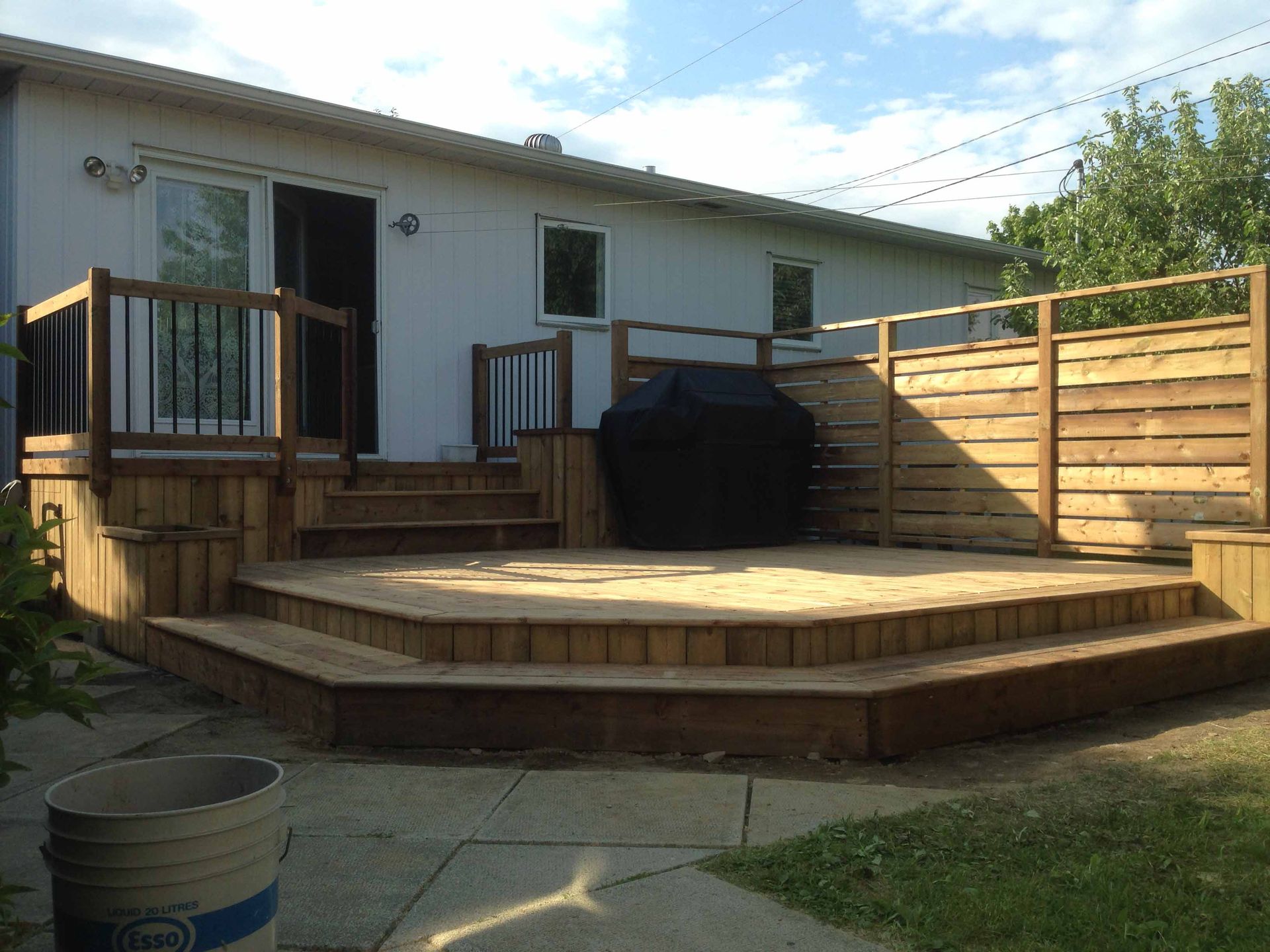 Terrasse arrière avec marches en bois incurvées, barbecue noir et revêtement extérieur de couleur claire.