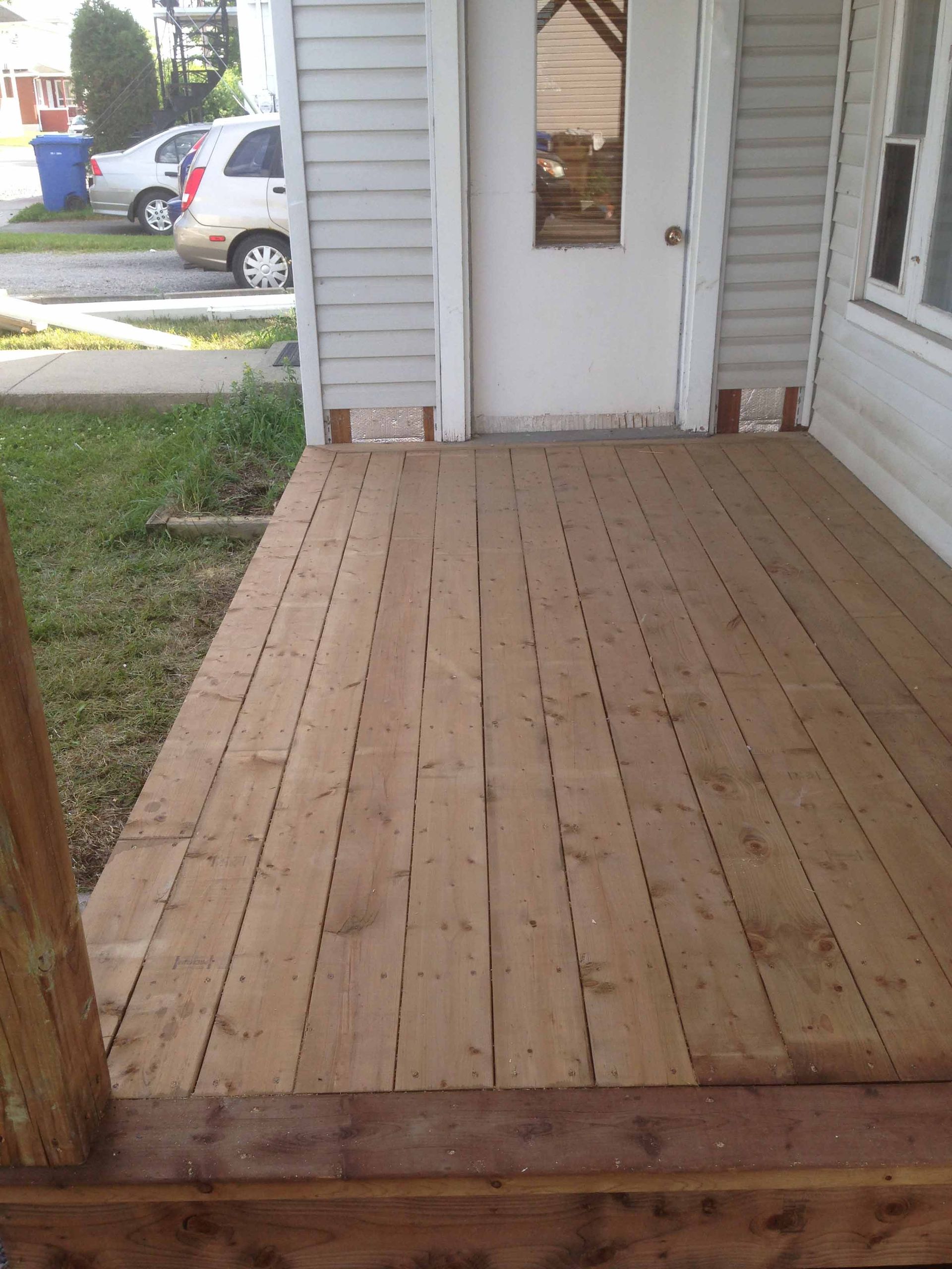 Une terrasse en bois mène à la porte d'entrée d'une maison blanche, avec une allée et des voitures garées à proximité.