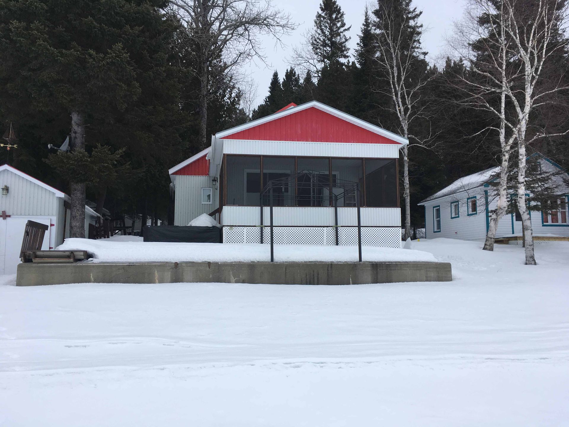 Maison enneigée au toit rouge et au porche, entourée d'arbres dans un paysage hivernal