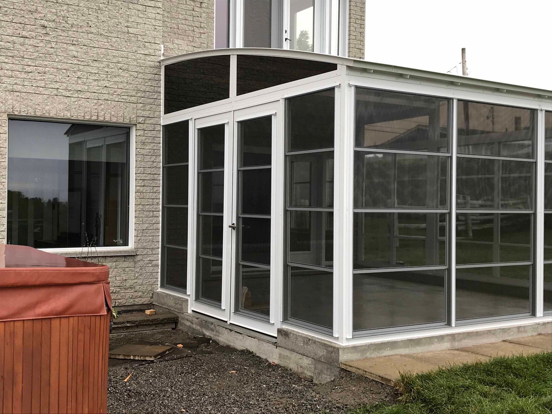 Véranda vitrée attenante à une maison en briques, avec une grande fenêtre et un abri pour jacuzzi à proximité.