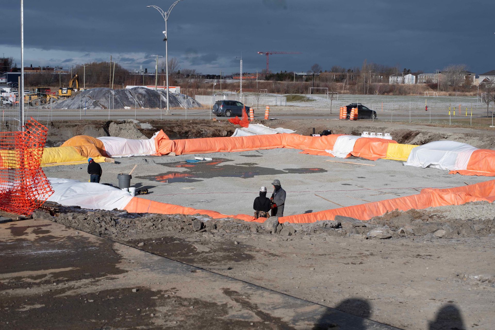 Chantier de construction avec filet de sécurité orange autour d'une grande excavation en béton sous un ciel nuageux
