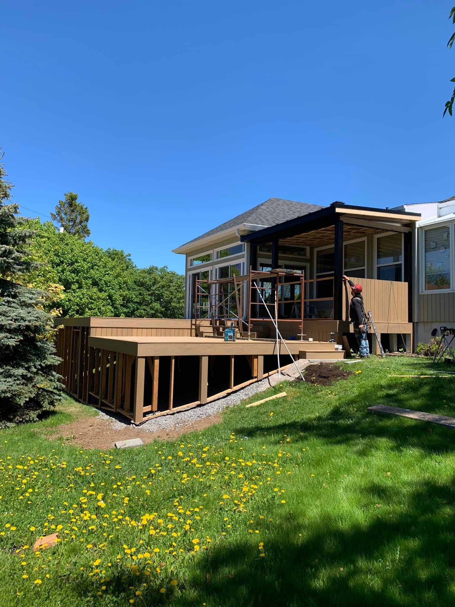 Terrasse arrière attenante à la maison, avec rambarde en bois, mobilier de jardin et pelouse verte sous un ciel bleu.