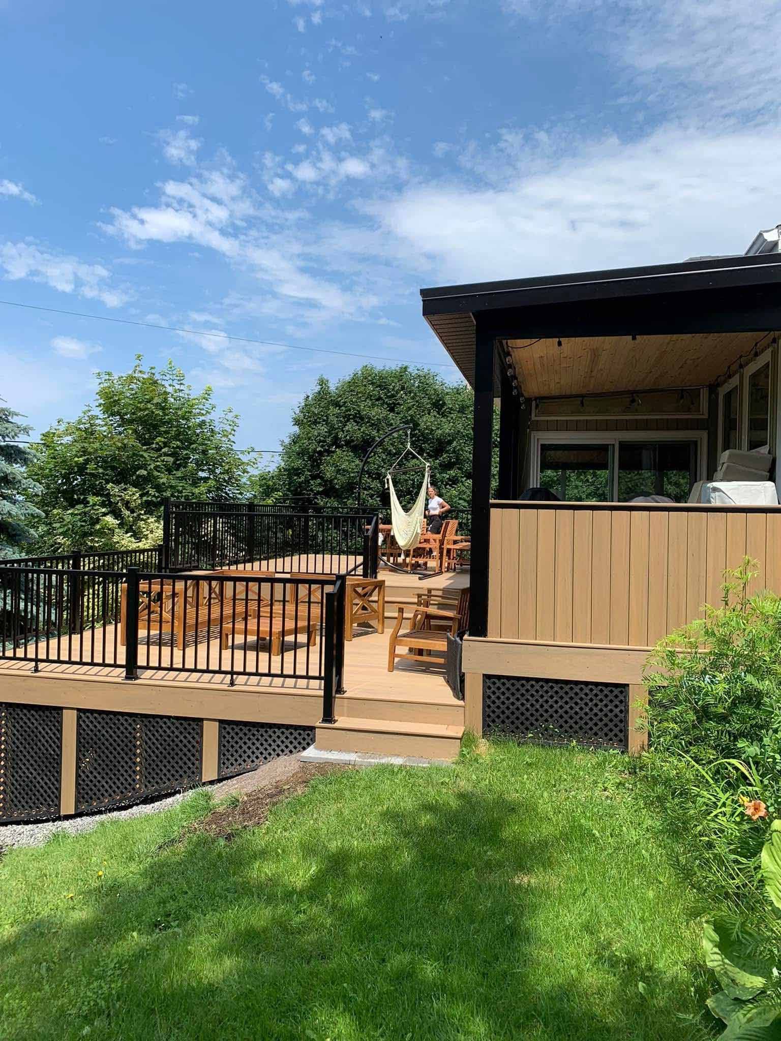 Terrasse et escalier en bois jouxtant une maison, donnant sur une cour gazonnée sous un ciel bleu.