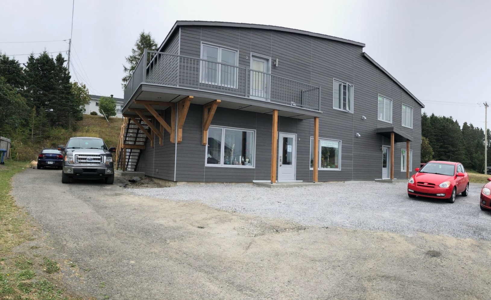 Maison grise à deux étages avec allée de gravier, SUV noir et voiture rouge garés, et véranda couverte