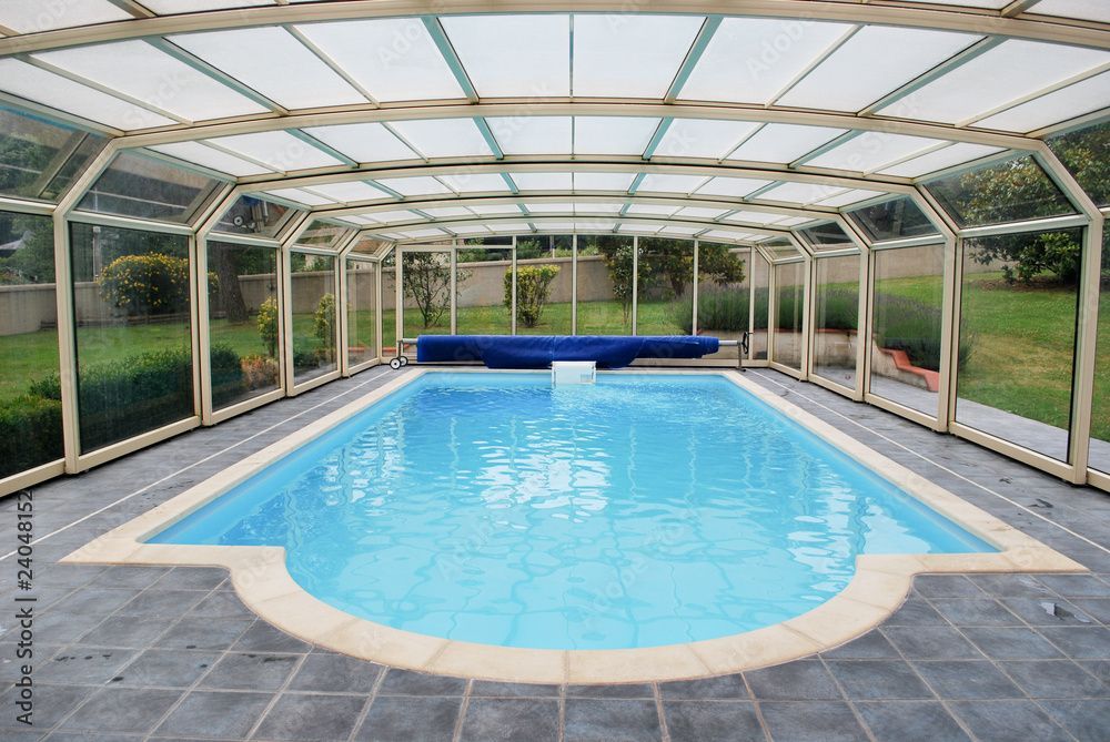 Piscine intérieure couverte avec eau bleue, terrasse carrelée et paroi transparente.