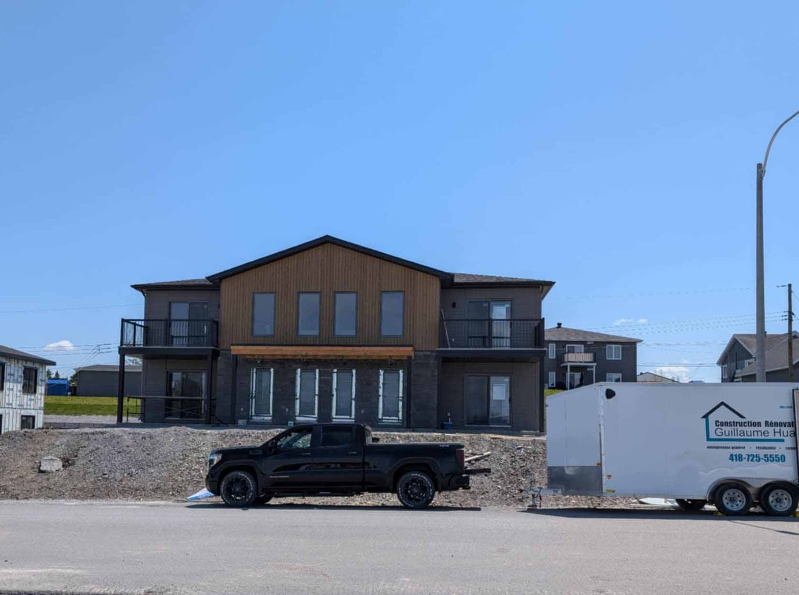 Maison brune de deux étages en construction, avec un pick-up noir et une remorque blanche devant.